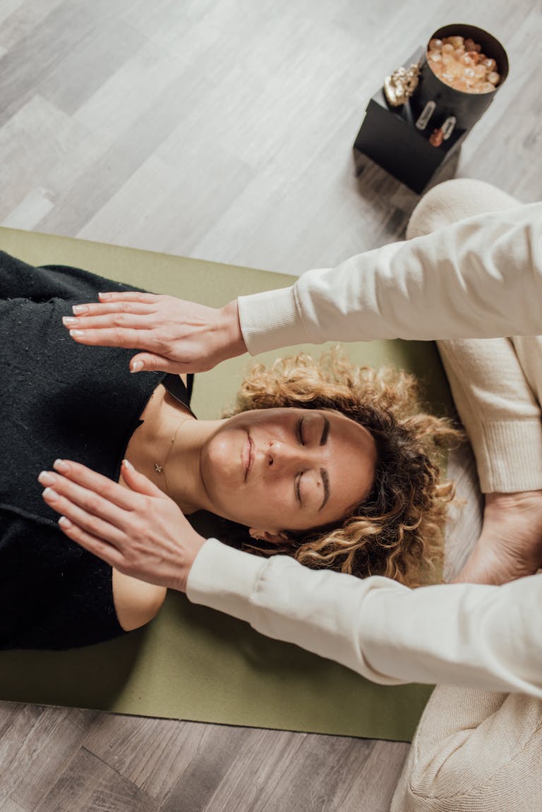 A woman receiving Reiki therapy with hands hovering over her, eyes closed, in a serene indoor setting.